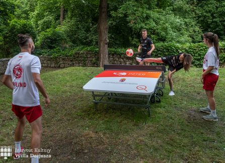 fotografia. dni wyszehradzkie w willi decjusza. mecz teqball na wolnym powietrzu. czworo zawodników wokół stołu ustawionego na trawie. w tle budynek willi decjusza.